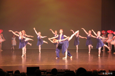 Bilde av en gruppe ballettdansere i blå kjoler på scenen. To dansere helt foran på scenen danser pas de deux. Det er en jente og en gutt som sammen danser i par. Gutten holder her jenta som lener seg framover med det eier beinet høyt opp bak.