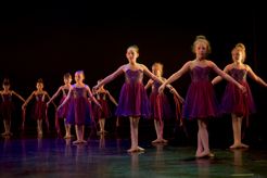 Bildet viser ni ballettdansere på scene. De har lilla kjole på og står i 3. posisjon med arner over skjørtene. 